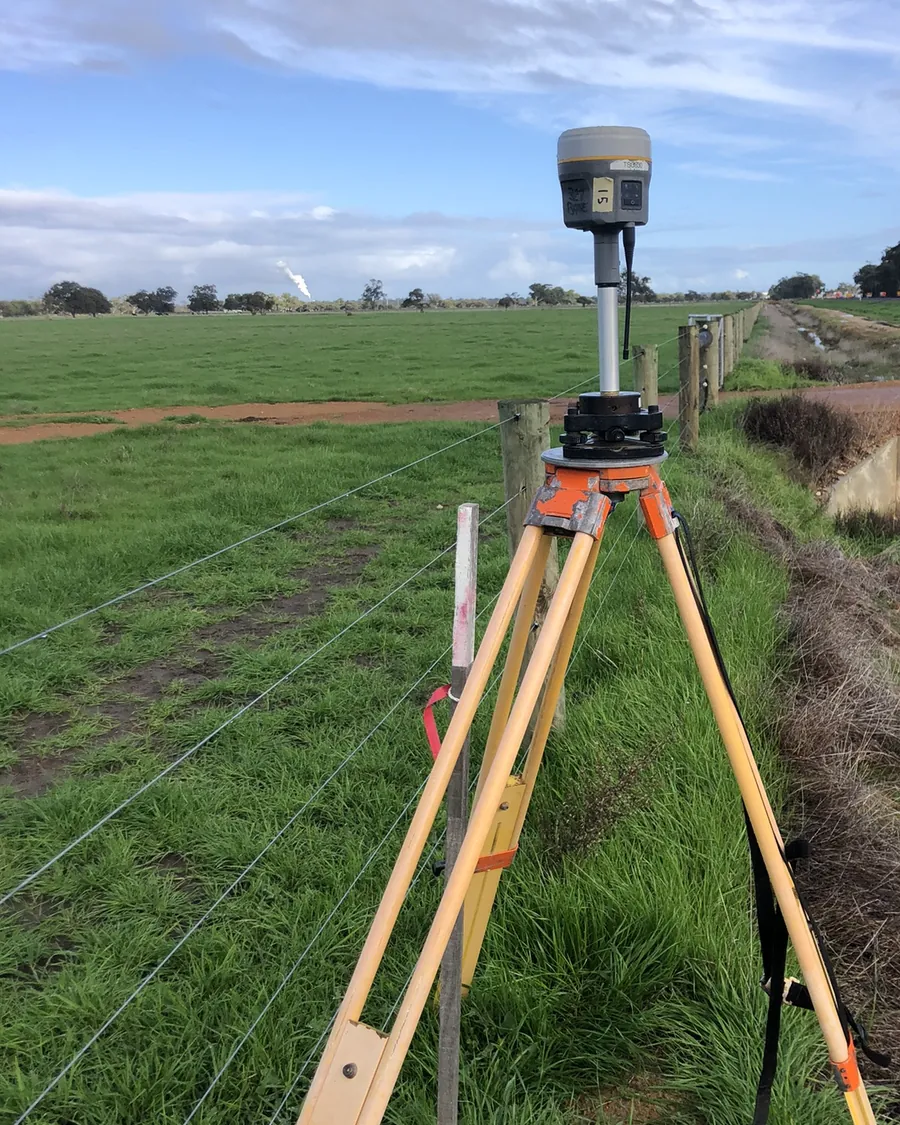 A GNSS receiver on a tripod set up over a survey mark beside a farm fence line